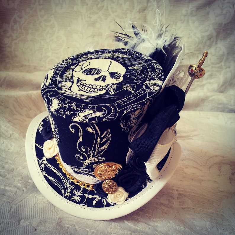 May include: A black and white top hat with a skull design, gold accents, and a white feather. The hat has a white ribbon and a white lace trim.