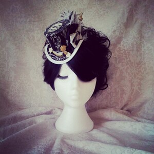 May include: A black and white mini top hat with gold accents and a feather. The hat is on a white mannequin head with black hair.