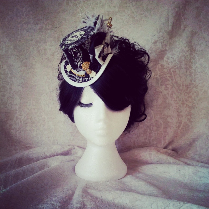 May include: A black and white mini top hat with gold accents and a feather. The hat is on a white mannequin head with black hair.