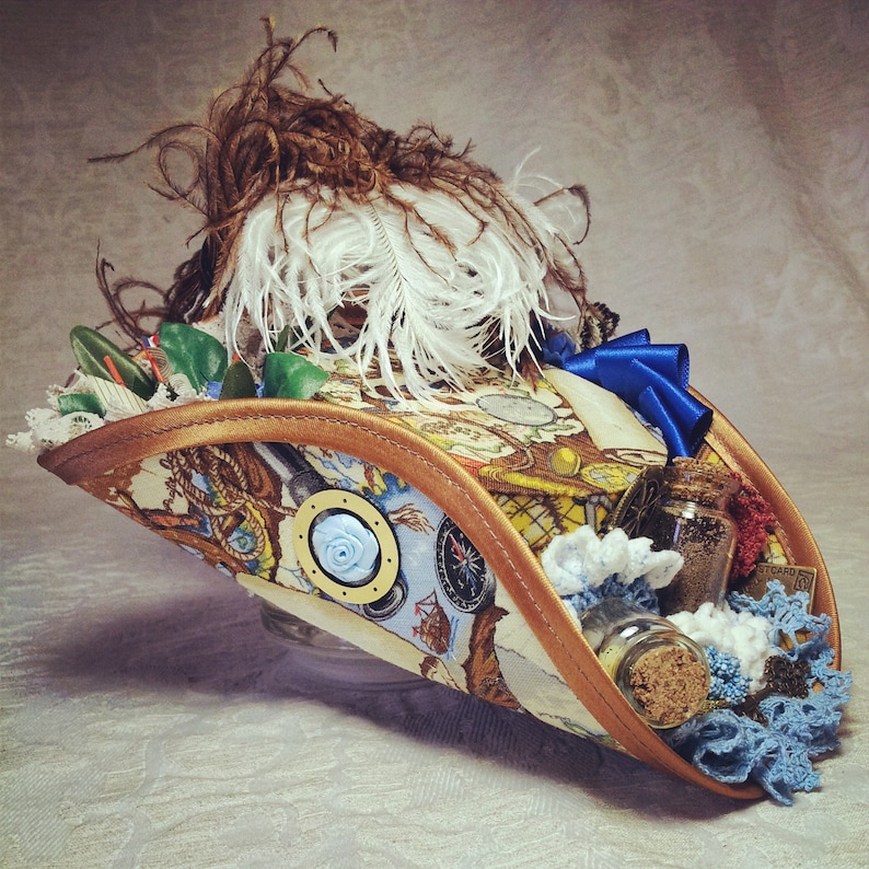 May include: A decorative pirate hat with a map-themed fabric, trimmed with brown and gold. It features white and brown feathers, a blue bow, and small glass bottles with corks. The hat is adorned with blue and white embellishments.