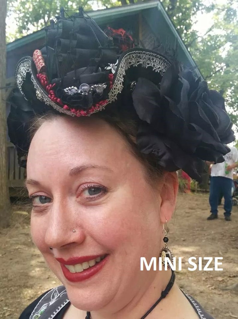 Puede incluir: Un sombrero negro y plateado con ribete rojo y blanco y una flor negra. El sombrero lo lleva una persona con un aro en la nariz y pendientes. El texto "MINI SIZE" es visible en la imagen.