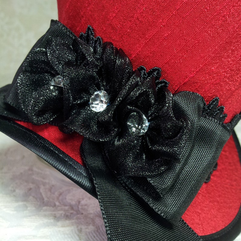 May include: A red top hat with a black ribbon and floral embellishments. The hat features a black ribbon band and is adorned with black fabric flowers and sparkling crystal accents. The hat is a stylish accessory.