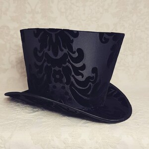 May include: A black top hat with a black damask pattern. The hat has a tall, cylindrical crown and a wide, flat brim.