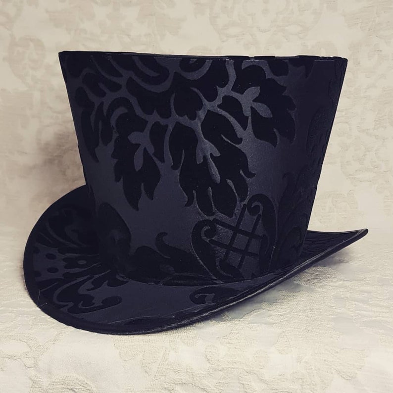 May include: A black top hat with a damask pattern. The hat has a wide brim and a tall crown.