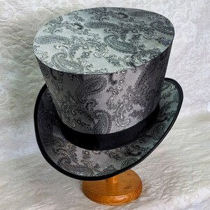 May include: A black and gray top hat with a paisley pattern. The hat is sitting on a wooden stand.