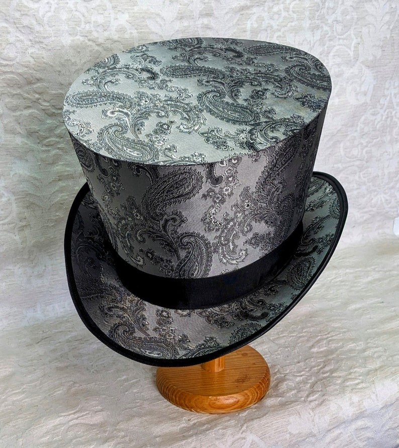 May include: A black and gray top hat with a paisley pattern. The hat is sitting on a wooden stand.