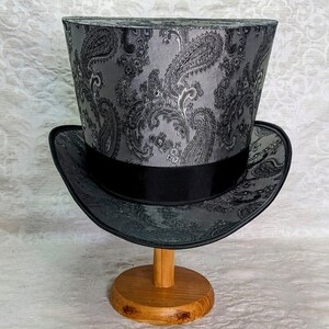 May include: A black top hat with a silver paisley pattern. The hat is sitting on a wooden stand.