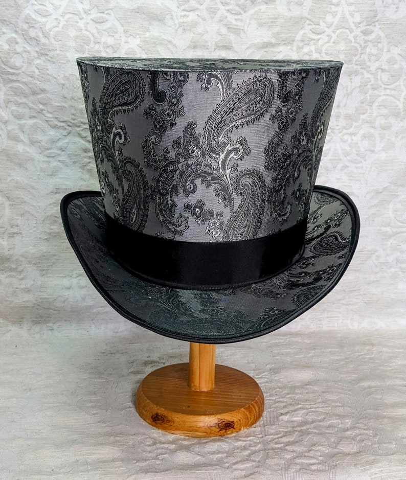 May include: A black top hat with a silver paisley pattern. The hat is sitting on a wooden stand.