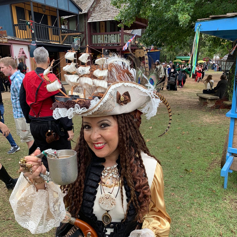 May include: A woman wearing a pirate-themed hat with a miniature ship on top, adorned with feathers and a brown bow. She is holding a metal mug, and wearing a gold and black outfit, with multiple necklaces and rings. The background shows a Renaissance fair setting.