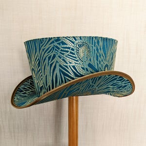 May include: A teal blue top hat with a gold and teal patterned design. The hat has a brown leather brim.