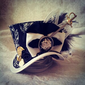 May include: A black and white top hat with a white bow and a gold and silver embellishment. A silver sword with a red jewel is attached to the hat.