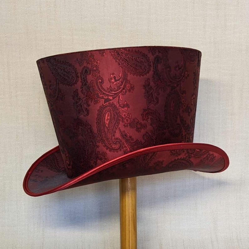 K&ouml;nnte beinhalten: Ein bordeauxfarbener Zylinderhut mit Paisley-Muster. Der Hut hat eine breite Krempe und eine hohe, zylindrische Krone. Der Rand und der Rand der Krone sind mit dunklerem rotem Satin verziert. Der Hut wird auf einem Holzst&auml;nder ausgestellt.