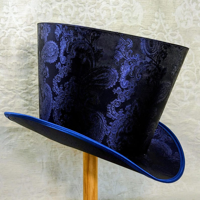 May include: A black top hat with a blue paisley pattern and a blue brim. The hat is made of a shiny fabric and is displayed on a wooden stand. The hat has a classic top hat shape.