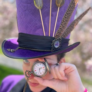 K&ouml;nnte beinhalten: Ein lila Zylinder mit schwarzem Band, verziert mit Federn, dekorativen Verzierungen und Lollipop-&auml;hnlichen Dekorationen. Eine Taschenuhr mit Kette wird vor das Auge gehalten. Der Hut hat ein gemustertes Design.