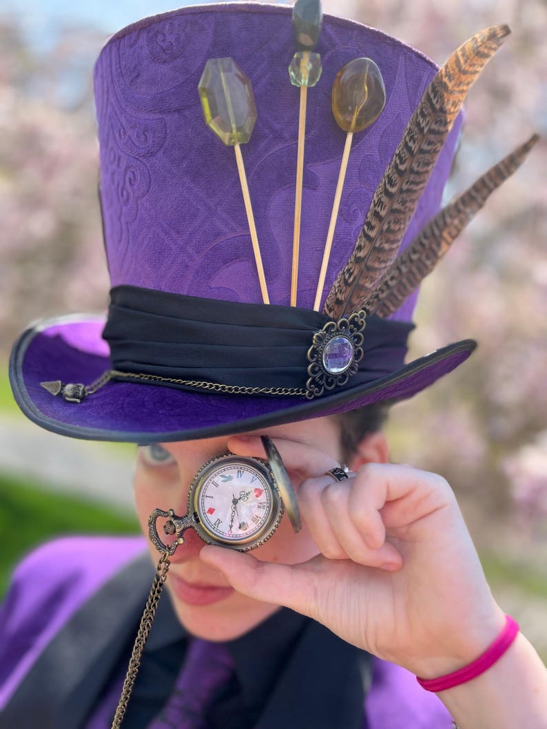 K&ouml;nnte beinhalten: Ein lila Zylinder mit schwarzem Band, verziert mit Federn, dekorativen Verzierungen und Lollipop-&auml;hnlichen Dekorationen. Eine Taschenuhr mit Kette wird vor das Auge gehalten. Der Hut hat ein gemustertes Design.