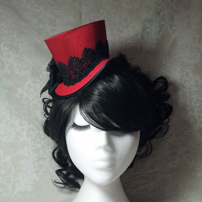 May include: A red mini top hat with black lace trim and a black bow. The hat is worn on a mannequin head with black hair. The hat's design is a mix of gothic and Victorian styles, suitable for cosplay or themed events.