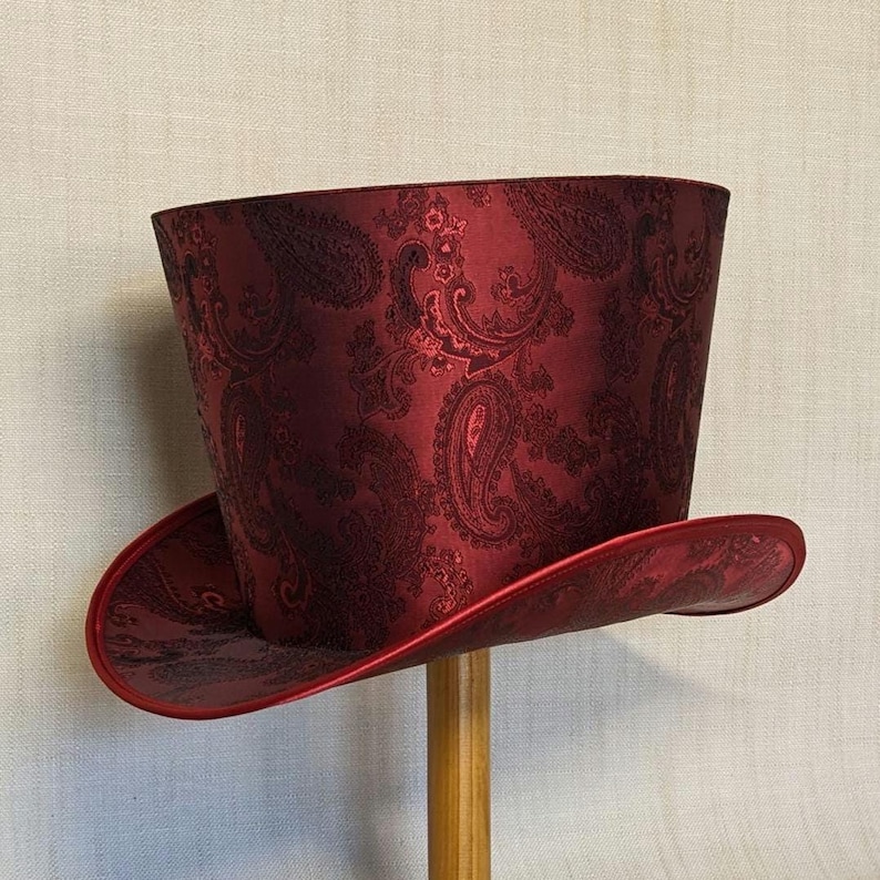 K&ouml;nnte beinhalten: Ein bordeauxfarbener Zylinderhut mit Paisley-Muster. Der Hut hat eine breite Krempe und eine hohe, zylindrische Krone. Der Hut ist aus einem gl&auml;nzenden Stoff gefertigt und auf einem Holzst&auml;nder montiert.