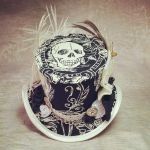 May include: A black and white top hat with a skull and floral design. The hat is decorated with feathers, lace, and beads.