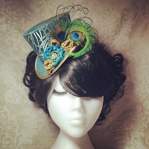 May include: A teal and gold top hat with a peacock feather and blue flower embellishment. The hat is worn on a mannequin with black hair.