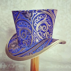 Puede incluir: Un sombrero de copa azul y dorado con un patrón de paisley. El sombrero tiene un ala ancha y una corona alta.