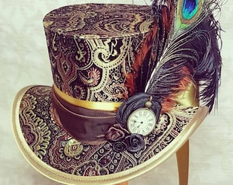 Puede incluir: Un sombrero de copa negro y dorado con un patrón de paisley y una pluma de pavo real. El sombrero tiene una cinta marrón y un pequeño reloj de bolsillo con números romanos.