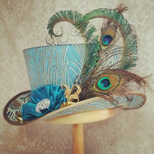 May include: A teal top hat with a floral design and peacock feathers. The hat features a teal satin flower with a rhinestone embellishment and a gold filigree accent. The brim is trimmed with gold and brown patterned fabric.