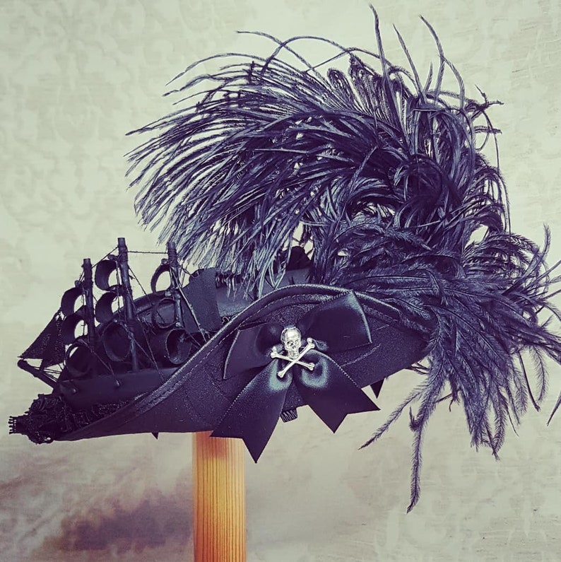 Puede incluir: Un sombrero de pirata negro con una pluma negra y un adorno de calavera y huesos cruzados. El sombrero tiene forma de tricornio y tiene un dise&ntilde;o de barco en la parte delantera.