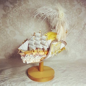 Puede incluir: Un sombrero decorativo blanco y dorado con un barco en miniatura y ribete de encaje. El sombrero est&aacute; adornado con plumas y un lazo amarillo.