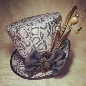 May include: A white and black snake skin patterned top hat with a black bow and a feather and bead embellishment.