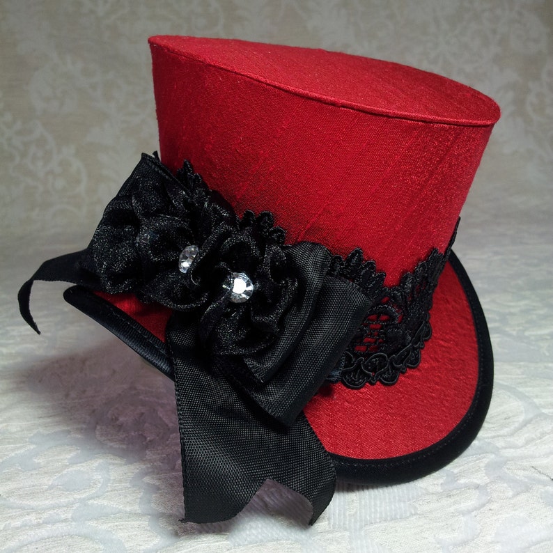 May include: A red mini top hat with a black ribbon and lace embellishments. The hat features a black flower with two sparkling gems and a black ribbon bow. The hat has a black brim and is likely for a costume or special event.