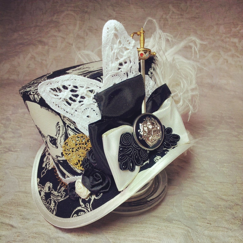 May include: A black and white top hat with a white lace trim, a black ribbon, and a gold and silver embellishment. A small sword is attached to the top of the hat.