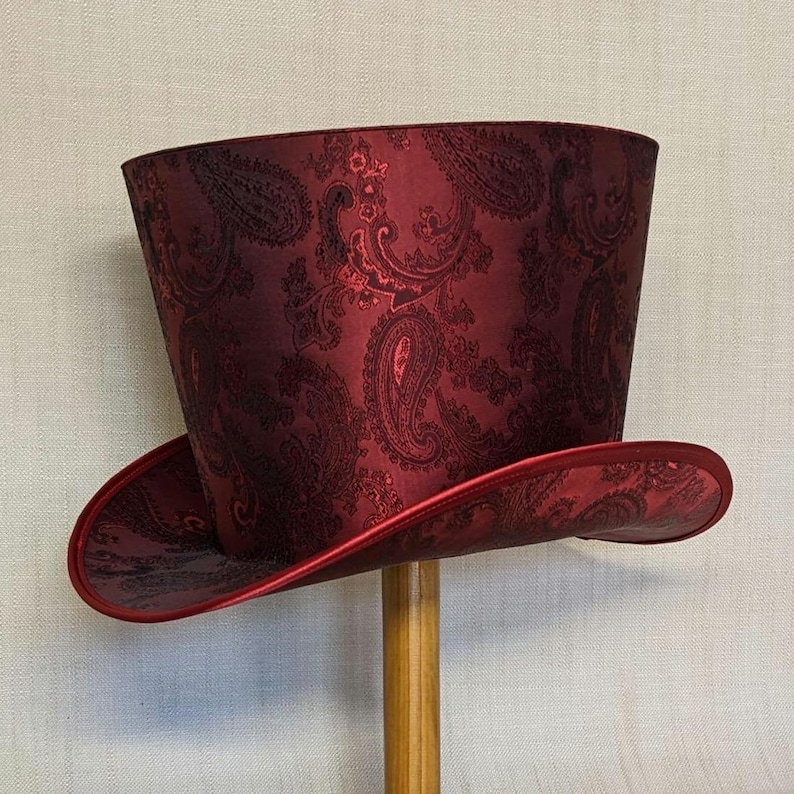 K&ouml;nnte beinhalten: Ein bordeauxfarbener Zylinderhut mit Paisley-Muster. Der Hut hat eine breite Krempe und eine hohe, zylindrische Krone. Der Stoff hat einen dezenten Glanz, und die Krempe ist mit passendem bordeauxfarbenem Stoff verziert. Der Hut wird auf einem Holzst&auml;nder pr&auml;sentiert.