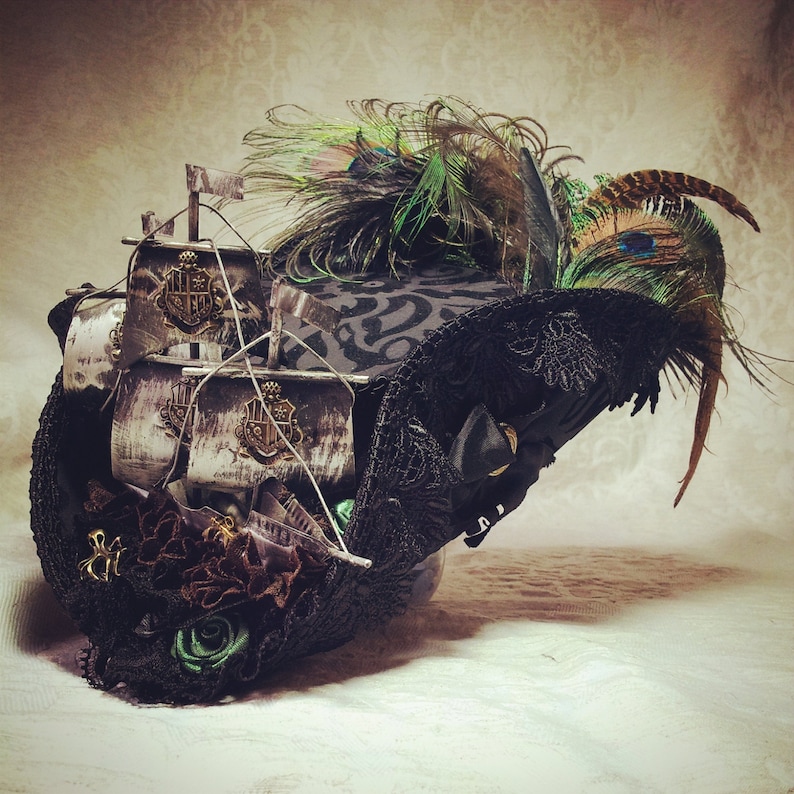 May include: A black steampunk-style hat with a miniature ship, peacock feathers, and ornate embellishments. The hat features a decorative ship with flags and crests, along with dark lace and floral accents. The design incorporates green and brown feathers.