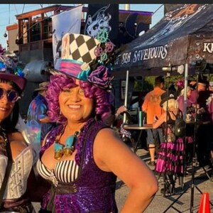 May include: A woman wearing a purple and black sequined vest with a white and black striped top underneath. She is wearing a large, colorful, and whimsical hat with a checkerboard pattern and a blue and white gear design. The hat has a purple feather and a blue flower. The woman has purple hair and is smiling. The background is blurry and shows a sign that says "Sea Saviours".