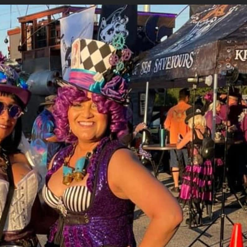 May include: A woman wearing a purple and black sequined vest with a white and black striped top underneath. She is wearing a large, colorful, and whimsical hat with a checkerboard pattern and a blue and white gear design. The hat has a purple feather and a blue flower. The woman has purple hair and is smiling. The background is blurry and shows a sign that says "Sea Saviours".
