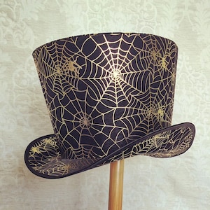 May include: A black top hat with a gold spiderweb pattern. The hat has a wide brim and a tall crown.