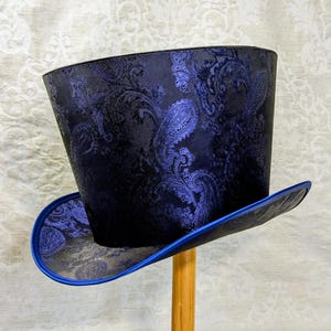 Op de afbeelding: Een marineblauwe hoge hoed met een paisley patroon in een lichtere blauwtint. De rand is afgewerkt met helderblauwe satijn. De hoed wordt op een houten standaard getoond.