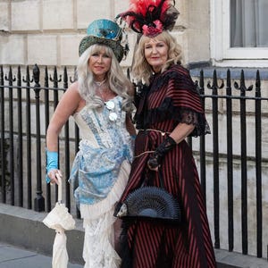 K&ouml;nnte beinhalten: Zwei Frauen in Vintage-Steampunk-Kleidung posieren im Freien. Eine tr&auml;gt ein blau-wei&szlig;es Korsettkleid mit passendem Hut und h&auml;lt einen wei&szlig;en Sonnenschirm. Die andere tr&auml;gt ein rot-schwarz gestreiftes Kleid mit Federhut und schwarzem F&auml;cher.