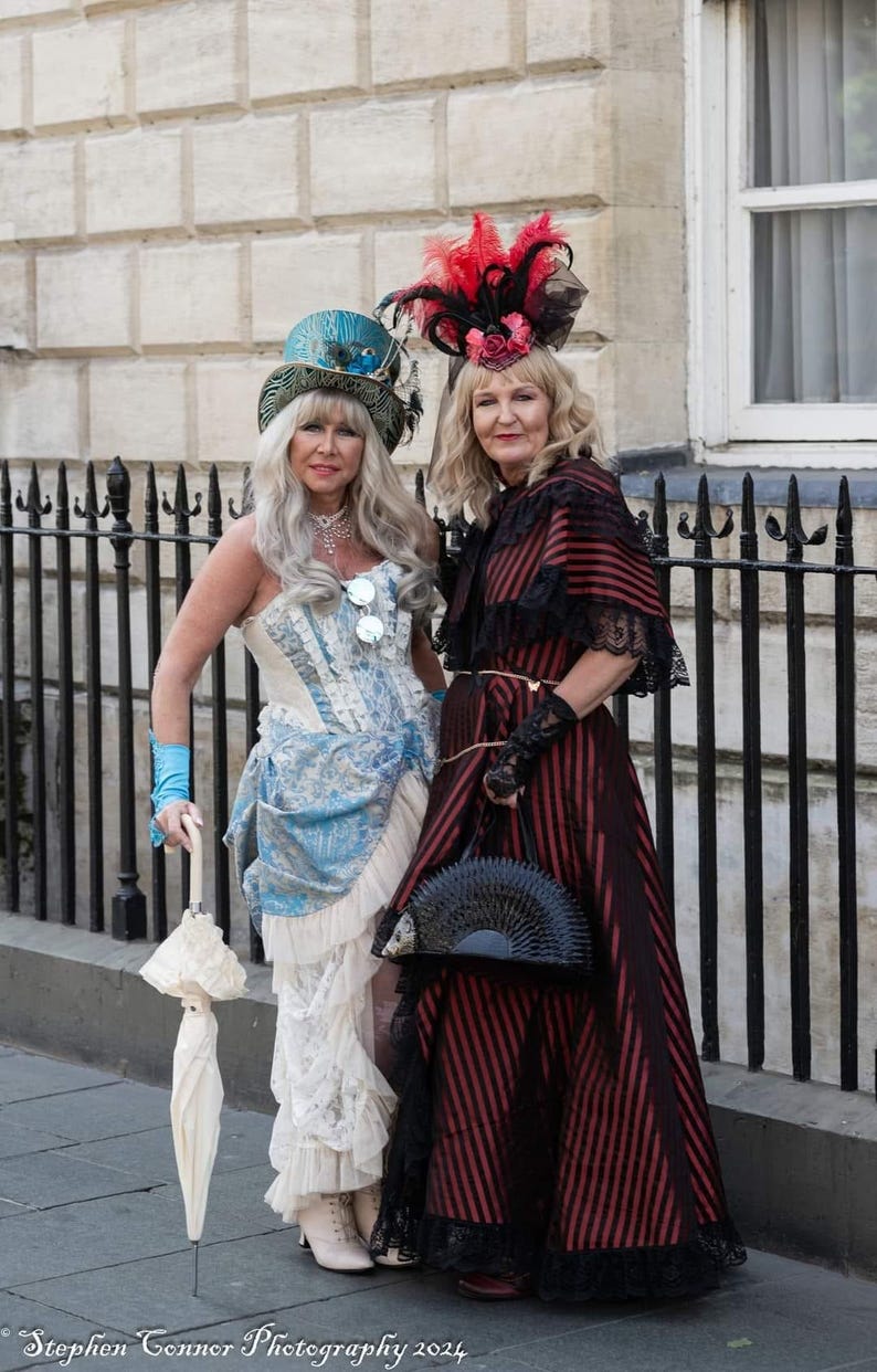 K&ouml;nnte beinhalten: Zwei Frauen in Vintage-Steampunk-Kleidung posieren im Freien. Eine tr&auml;gt ein blau-wei&szlig;es Korsettkleid mit passendem Hut und h&auml;lt einen wei&szlig;en Sonnenschirm. Die andere tr&auml;gt ein rot-schwarz gestreiftes Kleid mit Federhut und schwarzem F&auml;cher.