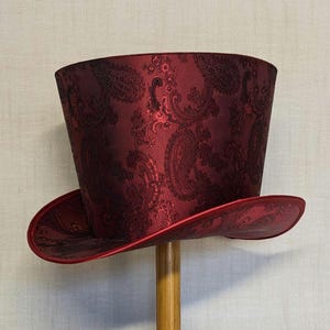K&ouml;nnte beinhalten: Ein bordeauxfarbener Zylinderhut mit Paisley-Muster. Der Hut hat eine breite Krempe und eine hohe, zylindrische Krone. Das Paisley-Design ist in einem dunkleren Burgunderrot gehalten, was einen subtilen Kontrast erzeugt. Der Hut wird auf einem Holzst&auml;nder pr&auml;sentiert.
