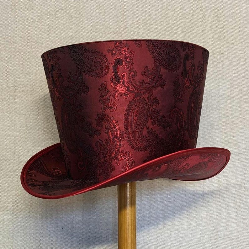 K&ouml;nnte beinhalten: Ein bordeauxfarbener Zylinderhut mit Paisley-Muster. Der Hut hat eine breite Krempe und eine hohe, zylindrische Krone. Das Paisley-Design ist in einem dunkleren Burgunderrot gehalten, was einen subtilen Kontrast erzeugt. Der Hut wird auf einem Holzst&auml;nder pr&auml;sentiert.