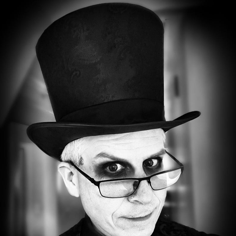 May include: Black and white portrait of a person wearing a black top hat and glasses. The person has white face makeup and dark eye makeup. The image is in black and white.