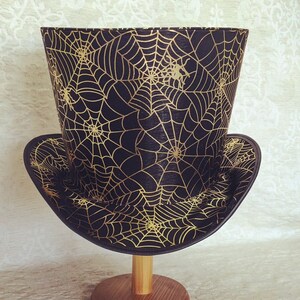 May include: A black top hat with a gold spiderweb pattern. The hat is on a wooden stand.