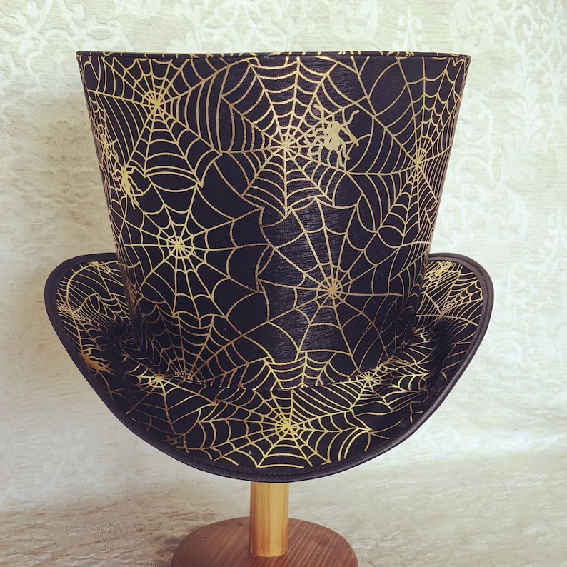 May include: A black top hat with a gold spiderweb pattern. The hat is on a wooden stand.