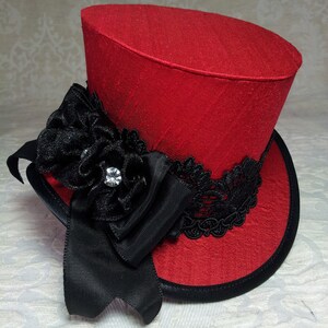 May include: A red top hat with a black ribbon, lace, and a decorative flower with a rhinestone. The hat has a black brim and a textured fabric. This miniature hat is a unique accessory.