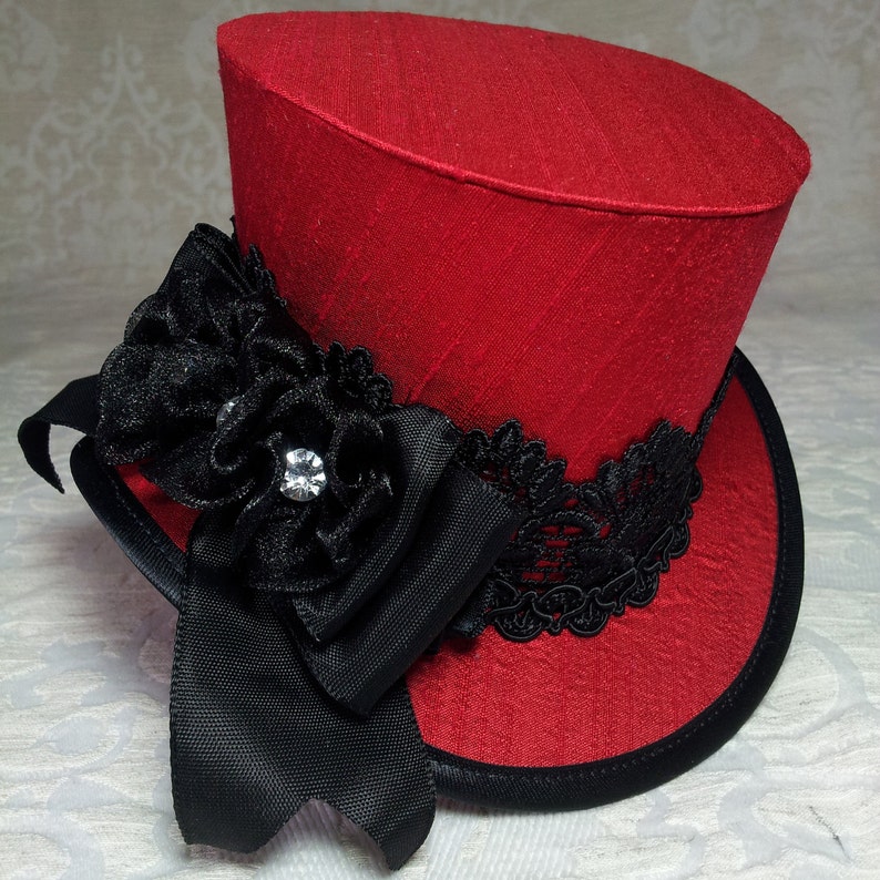 May include: A red top hat with a black ribbon, lace, and a decorative flower with a rhinestone. The hat has a black brim and a textured fabric. This miniature hat is a unique accessory.