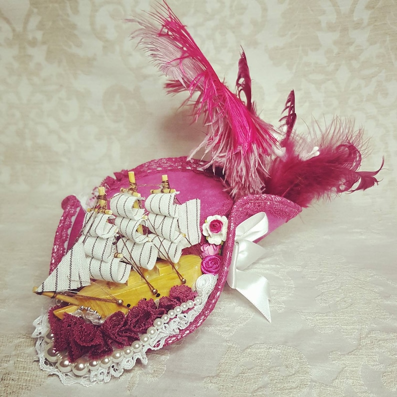 K&ouml;nnte beinhalten: Ein rosa dekorativer Hut mit einem Miniaturschiff, wei&szlig;er Spitze und Perlenakzenten. Der Hut hat eine gro&szlig;e rosa Feder und eine wei&szlig;e Satinschleife. Das Schiff hat wei&szlig;-grau gestreifte Segel.