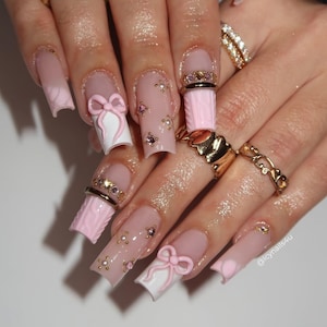 May include: Long, square-shaped acrylic nails with a light pink ombre design. Some nails feature 3D white bows, while others have small gems and pearls. Gold rings adorn the fingers.