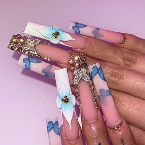 May include: Long, square-shaped acrylic nails with a light pink and white ombre design. Some nails feature 3D light blue flowers with gold accents, while others have blue butterfly designs. Several nails are adorned with gold rhinestones.
