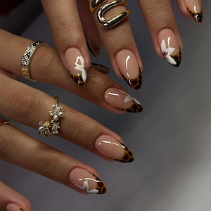 May include: Close-up of manicured nails with a nude base, brown tortoise shell tips, and white floral accents. The nails are long and almond-shaped. Gold rings adorn the fingers.